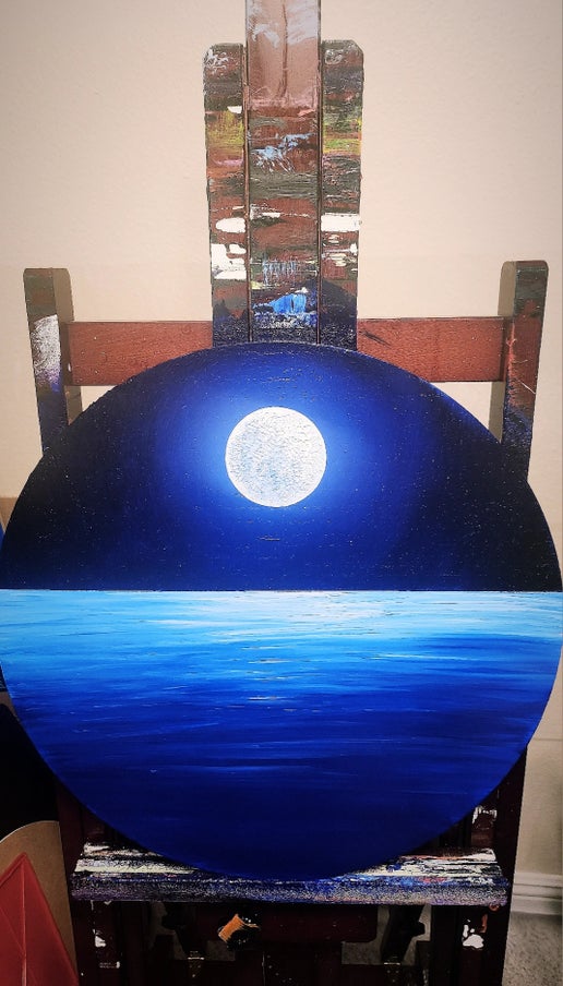 Wooden circle painting of a bright full moon and blue ocean sitting on an easel