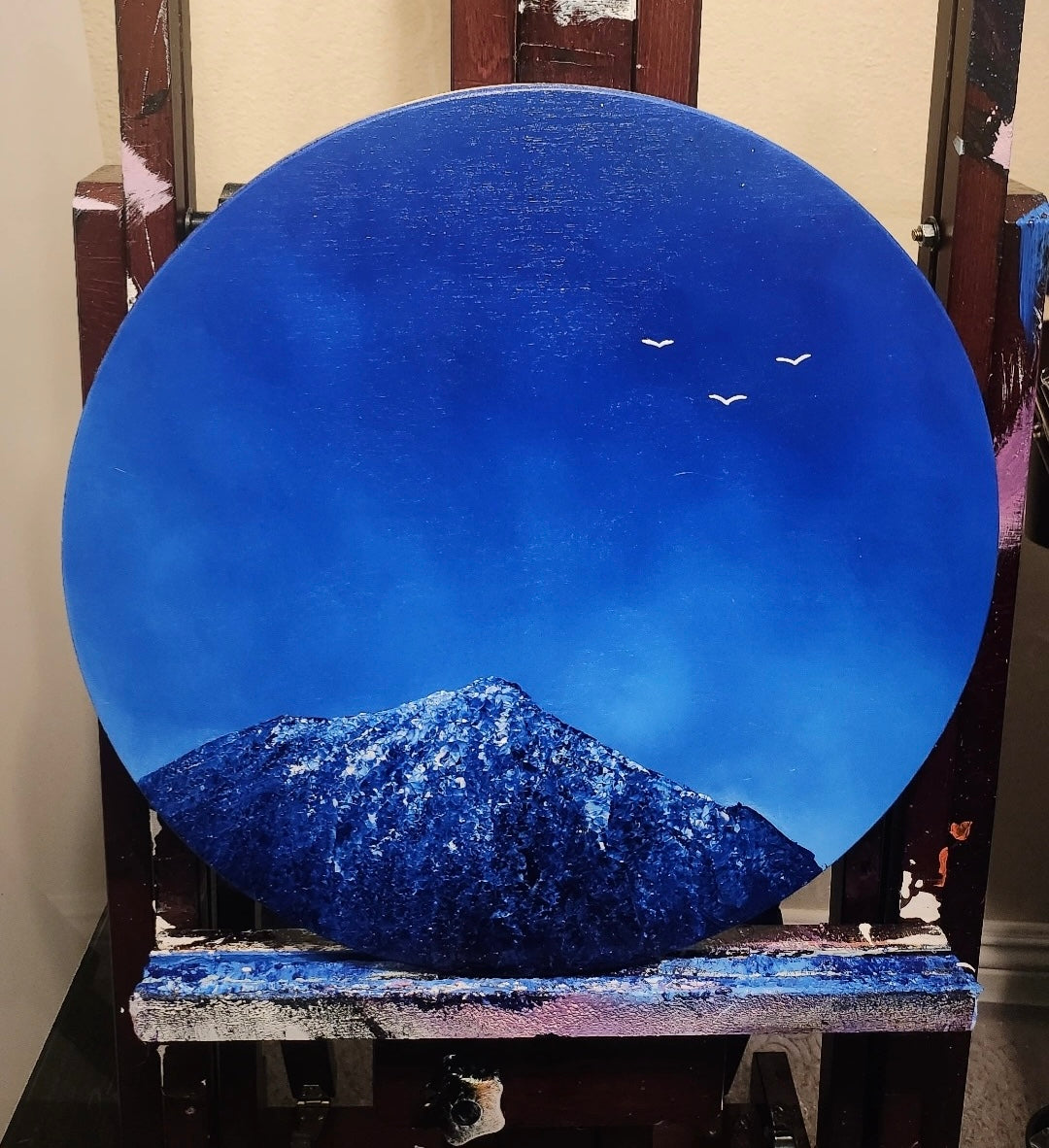 Round blue painting of a mountain and sky with birds on an easel