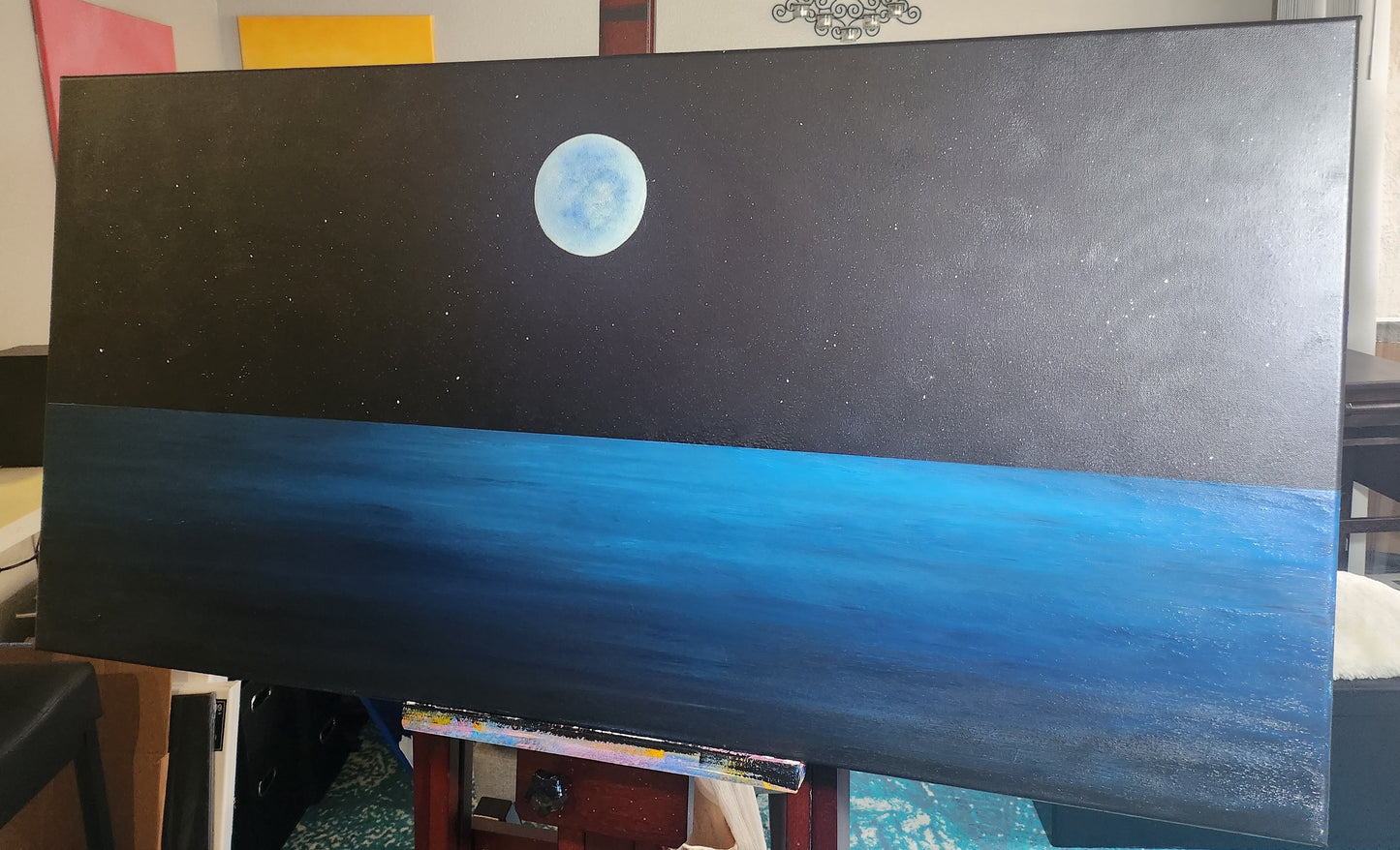 A painting on an easel of a dark night sky above a blue ocean and a full moon