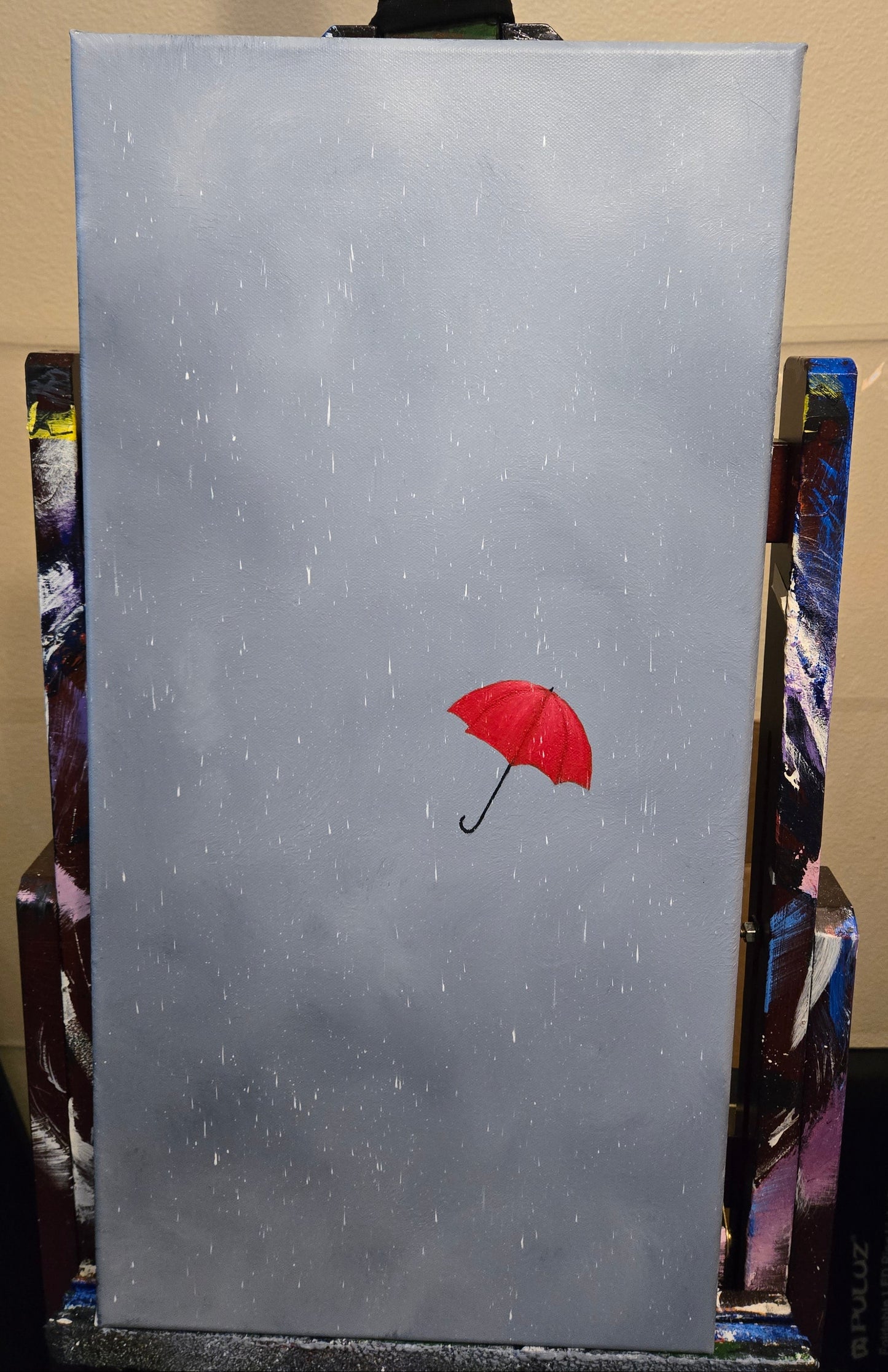 A painting on an easel of a red umbrella against a rainy grey cloudy storm