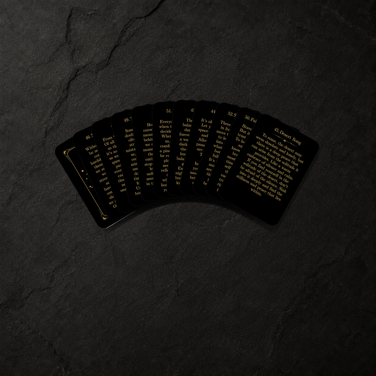 A fan of black oracle cards with gold text, displayed on a dark textured slate background.