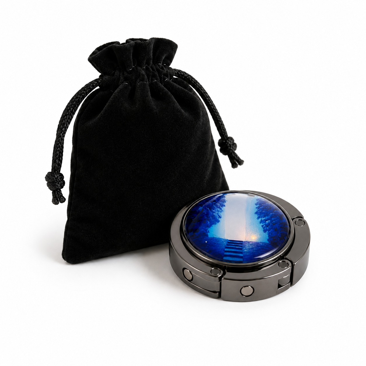 A round metal bag hook with artwork on the front sitting in front of a small black drawstring pouch on a clean white background.