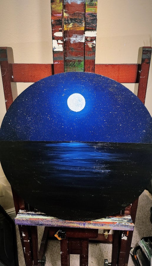 Round painting of a night sky with a full moon over water, displayed on a stand.