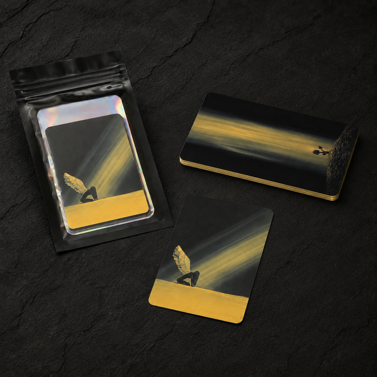 A stack of cards showing gold gilding on the side and an example card with a black and gold desert landscape design are arranged on a dark textured surface, including all the cards sealed in a transparent holographic pouch.