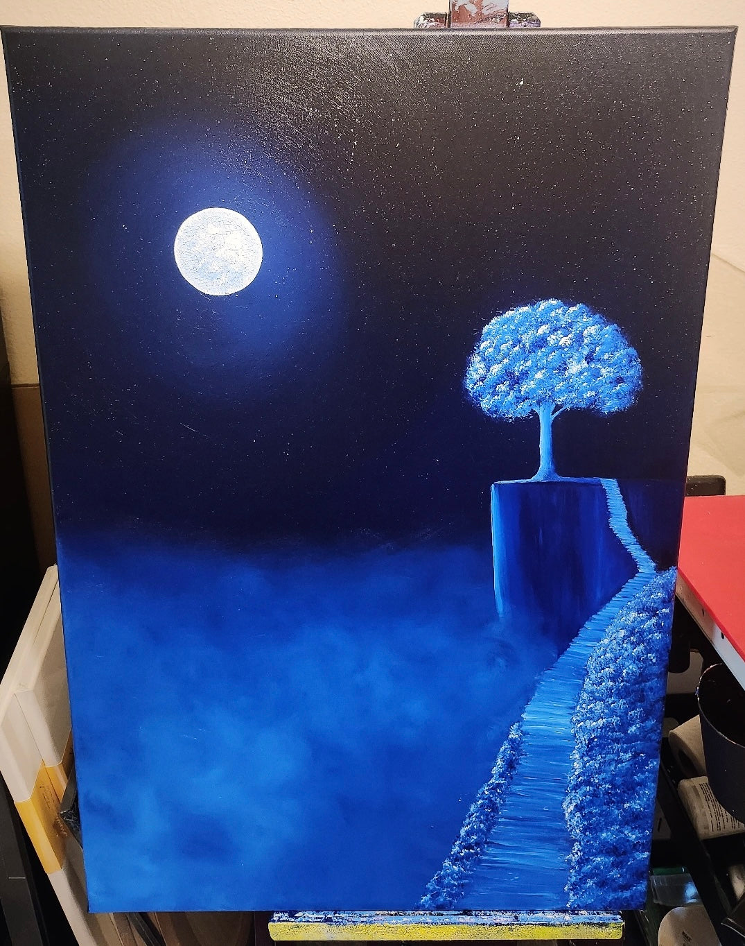 Painting on an easel of a blue tree on a ledge with a full moon in the night sky.